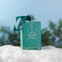 An UpCircle Winter Glow Duo box in green, tied with a ribbon, sits on snow amid falling snowflakes and blurred pine trees under a clear blue sky—the perfect sustainable gift.