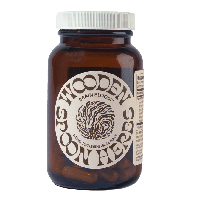 A brown glass bottle labeled Brain Bloom Capsules by Wooden Spoon Herbs holds 60 capsules. It features a round beige label with wavy text, a brain illustration, and contains nootropic herbs to support cognitive function.