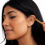A woman with long dark hair and multiple ear piercings has a WTHN Ear Seed Kit gold tweezer gently placed on her ear, suggesting the use of ancient Chinese Medicine techniques for pain relief, against a plain white background.