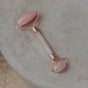 A pink jade WTHN Face Roller Kit with rose gold accents lies on a textured gray surface. Inspired by Traditional Chinese Medicine, it features two polished stone ends to reduce puffiness. WITH is printed on the handle.