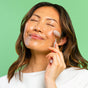 A woman with medium-length brown hair, eyes closed and relaxed, uses the WTHN Face Roller Kit—a tool inspired by Traditional Chinese Medicine—gently massaging her cheek against a green background while wearing a white top.