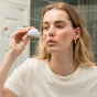 A blonde woman wearing gold jewelry uses the WTHN Face Cupping Kit for an all-natural under-eye lift while standing in her bathroom and looking at her reflection.