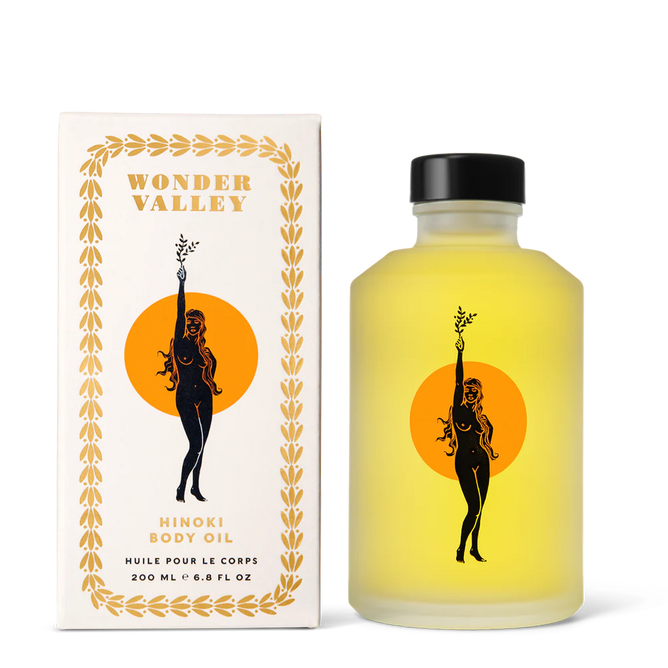 A yellow bottle of Wonder Valley Hinoki Body Oil with a black cap stands by its box. Both show a woman with a branch, an orange circle behind her, and decorative borders. Text: 