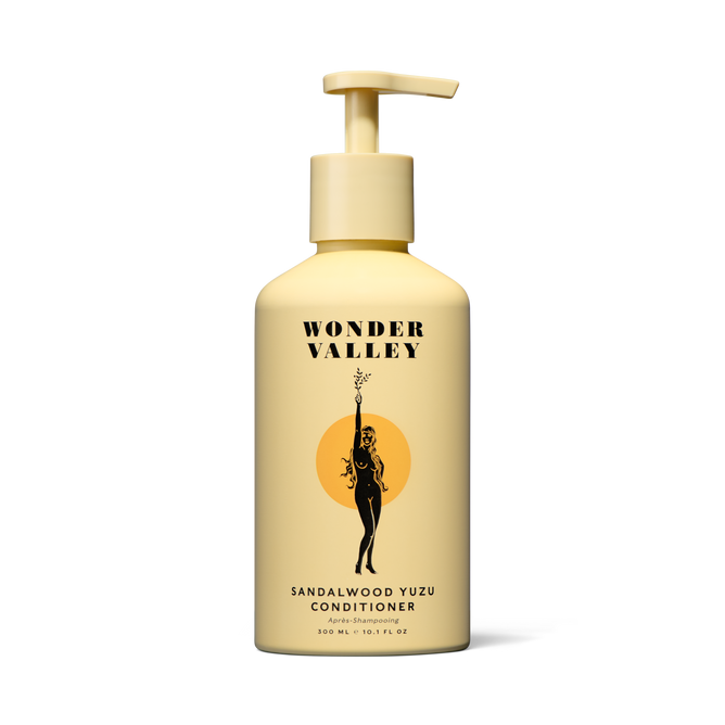 A yellow pump-top bottle of Wonder Valley Sandalwood Yuzu Conditioner with black text and a woman-branch illustration on an orange circle, formulated to reduce breakage and boost healthy shine.