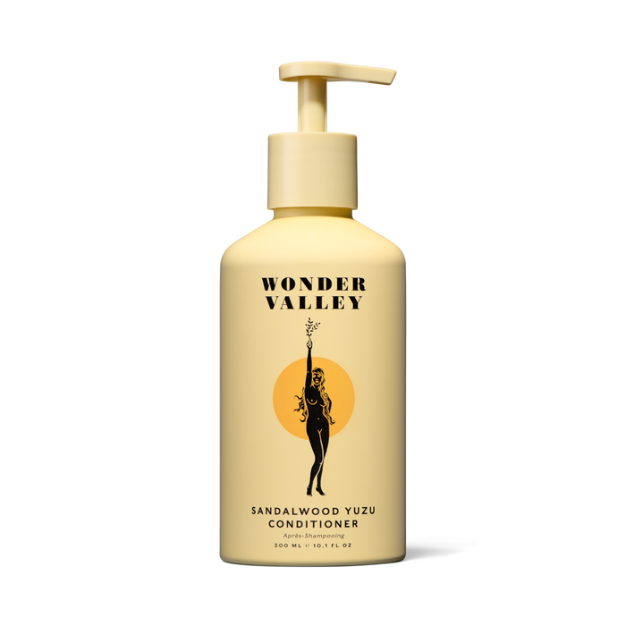 A yellow pump-top bottle of Wonder Valley Sandalwood Yuzu Conditioner with black text and a woman-branch illustration on an orange circle, formulated to reduce breakage and boost healthy shine.
