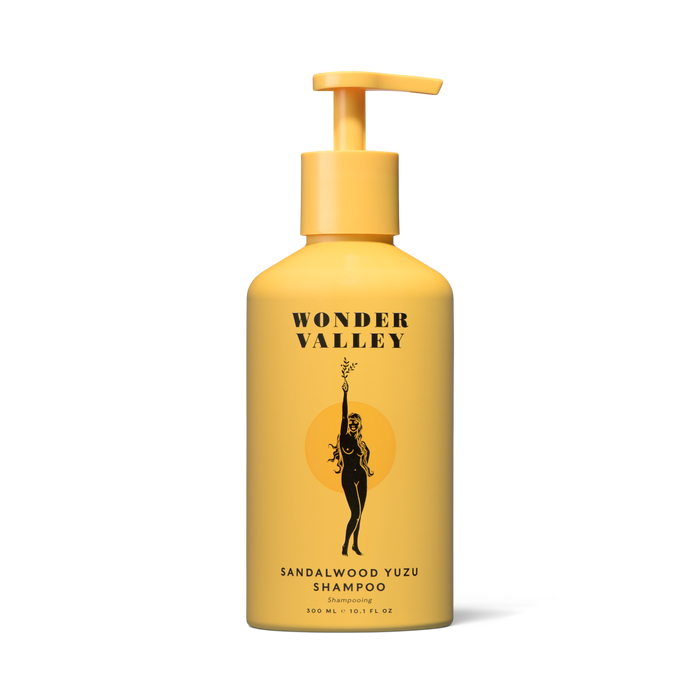 A yellow sustainable aluminum bottle of Wonder Valley Sandalwood Yuzu Shampoo, featuring an illustration of a woman holding a branch, is shown against a plain white background.