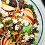 A fresh salad in a glass bowl with mixed greens, sliced red apples, walnuts, red onion, crumbled feta cheese, and a drizzle of Kosterina Walnut Balsamic Vinegar inspired by Modena, Italy.
