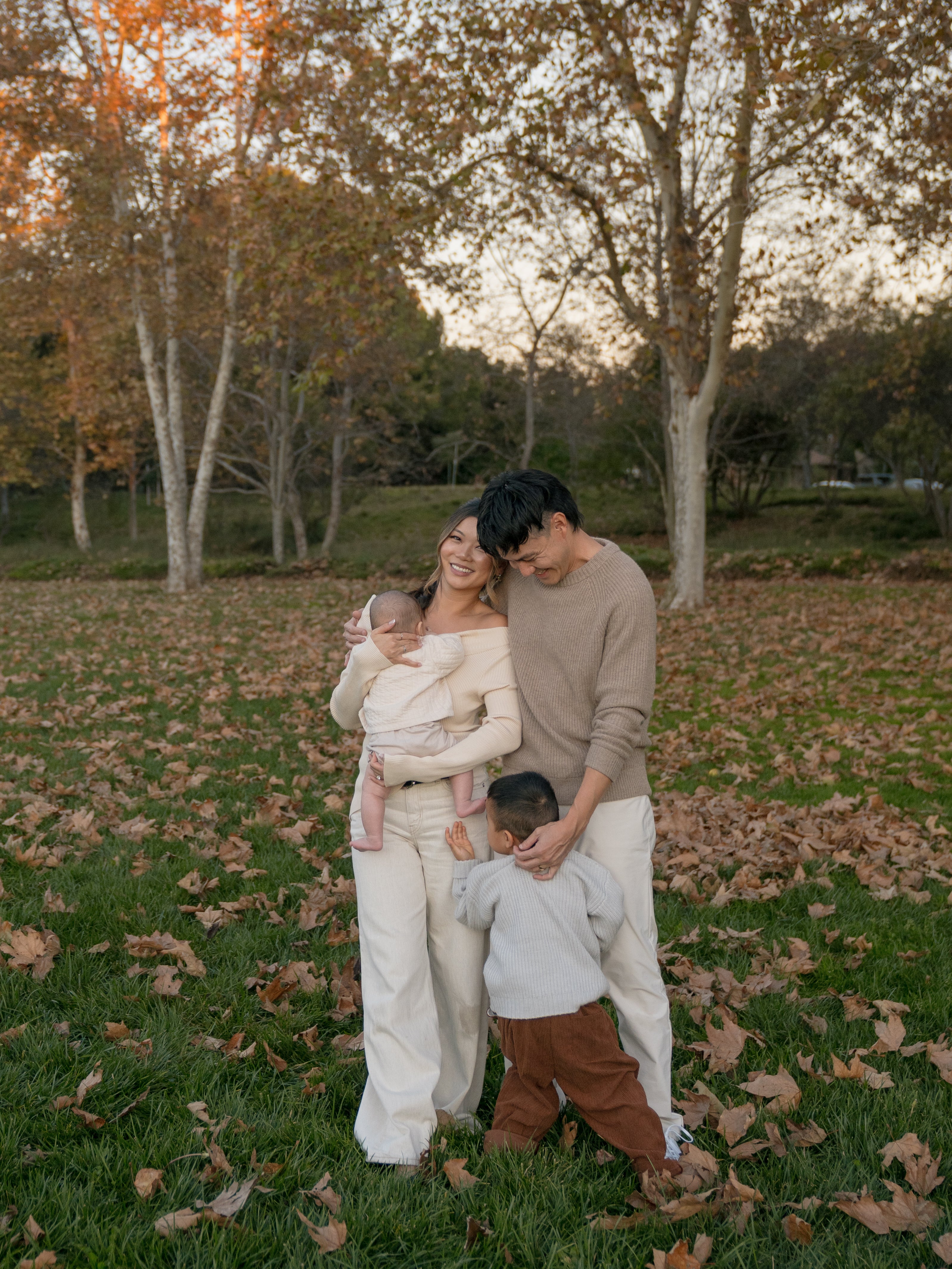 A smiling family of four stands outdoors on grass with fallen leaves. The mother holds an infant, the father stands beside her, and a young child hugs the fathers leg. Trees with autumn leaves are in the background.