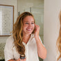 A woman with long wavy hair smiles while applying Chagrin Valley Soap & Salve’s Whipped Squalane Face & Eye Mousse in front of a mirror. She wears a white shirt and holds the cream jar, with framed art in the background.