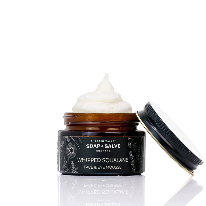 An open jar of Chagrin Valley Soap & Salve Whipped Squalane Face & Eye Mousee sits on a white background, its creamy mousse filling the jar. The black floral label is visible, with the lid placed next to the plant-powered moisturizer.