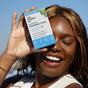 A smiling woman holds a box of The Humble Co. Advanced Whitening Strips near her face, displaying her white teeth outdoors—thanks to their advanced formula.