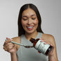 A smiling woman with long brown hair and a sleeveless top pours Davids Hydroxi™ Oil Pulling Oral Rinse with Nano Hydroxyapatite from a brown bottle onto a spoon, preparing for an oral care routine that supports fresh breath and enamel health.