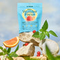 A brightly lit, nature-inspired scene features a blue pouch of Plant People WonderFocus - Mushroom Gummies with Lion’s Mane, surrounded by rocks, greenery, sliced grapefruit, white mushrooms, and scattered red nootropic gummies.