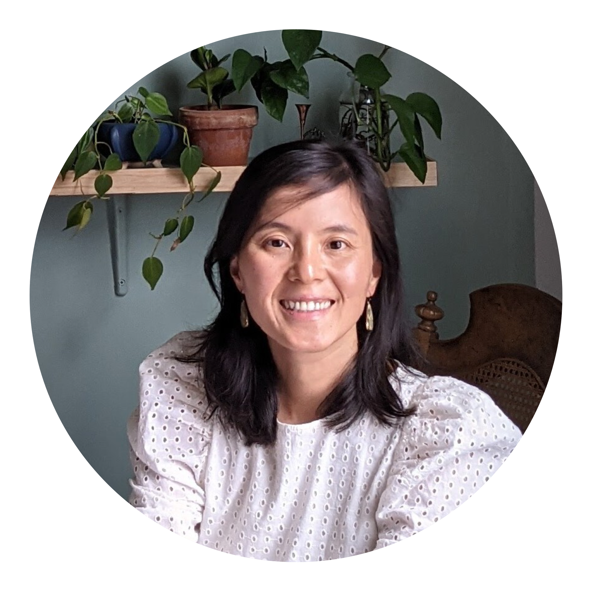 A woman with shoulder-length dark hair, wearing a white eyelet blouse, sits and smiles in front of a shelf with potted plants against a muted green wall.