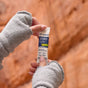 Close-up of hands in gray fingerless gloves holding a packet of Trace Minerals ZeroLyte, an electrolyte and sodium drink mix for hydration and muscle support, against a blurred reddish-brown rocky background.