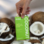 A hand holds a green Zimba Whitening Strips box above opened coconuts and coconut pieces—ideal for anyone wanting whiter teeth while reducing sensitivity.