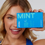 A smiling woman holds a blue Zimba Whitening Strips box labeled MINT and teeth whitening strips near her face, displaying her white teeth. The box advertises 14 treatments for professional whitening results.