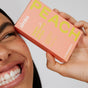 A person grins, displaying whiter teeth, while holding a peach box labeled Zimba Whitening Strips near their face. The box highlights 14 treatments of effective whitening strips from Zimba.