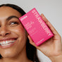 A person with clear skin smiles while holding a pink box of Zimba Whitening Strips close to their face, highlighting their whiter teeth.