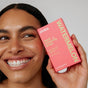 A smiling person holds a bright pink box of Zimba Whitening Strips near their face. The bold packaging displays white and yellow text and contains 14 strips for a visibly whiter smile.