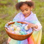 A young girl in a pastel rainbow dress and cape holds a basket of delicate flowers and Reusable and Fillable Wooden Easter Eggs from Sarahs Silks, crafted from FSC-certified wood. Wrapped in colorful cloths, shes outdoors on grass, gazing gently at the eggs.