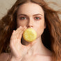 A person with long, wavy auburn hair prone to frizz holds The Balancer Shampoo Bar by NOT!CE Hair Co in front of their mouth, partially concealing it. Their neutral expression and freckles against a plain background capture the essence of plastic-free hair care.