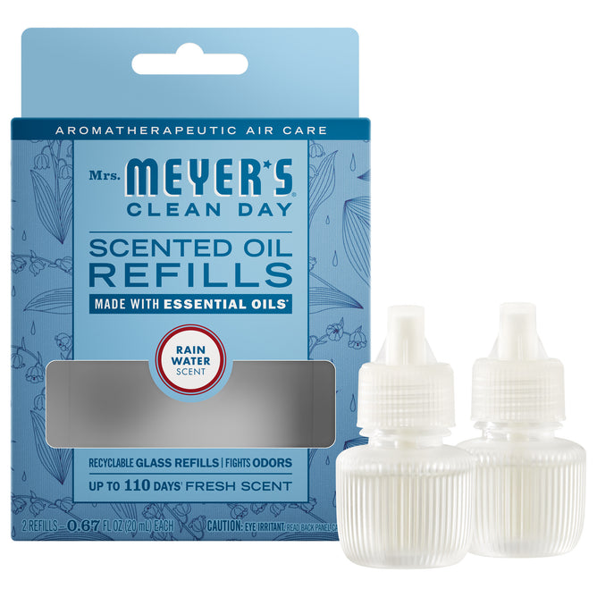 A blue box of Mrs. Meyer’s Scented Oil Refills 2pk sits next to two white diffuser bottles with droppers. The box features product info, floral design, and promotes garden-inspired scents.