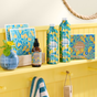 A yellow shelf displays Grove Co.s limited edition spring collection Laundry Sheets, featuring floral designs. Two spray bottles, a cleaner box, and a smaller bottle complete the setup. A small plant, cloth, and rope accent add charm to the arrangement.