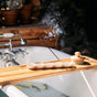 A lit incense stick in a ceramic holder emits smoke on a wooden bath tray beside a dry brush. A bathtub filled with water and floating lemon slices is in the background.