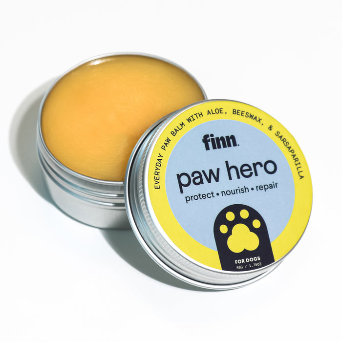 A tin of Finn Paw Hero balm for dogs, with the lid partially open to show the yellow balm inside. The label highlights ingredients like aloe and beeswax and states it protects, nourishes, and repairs paws.
