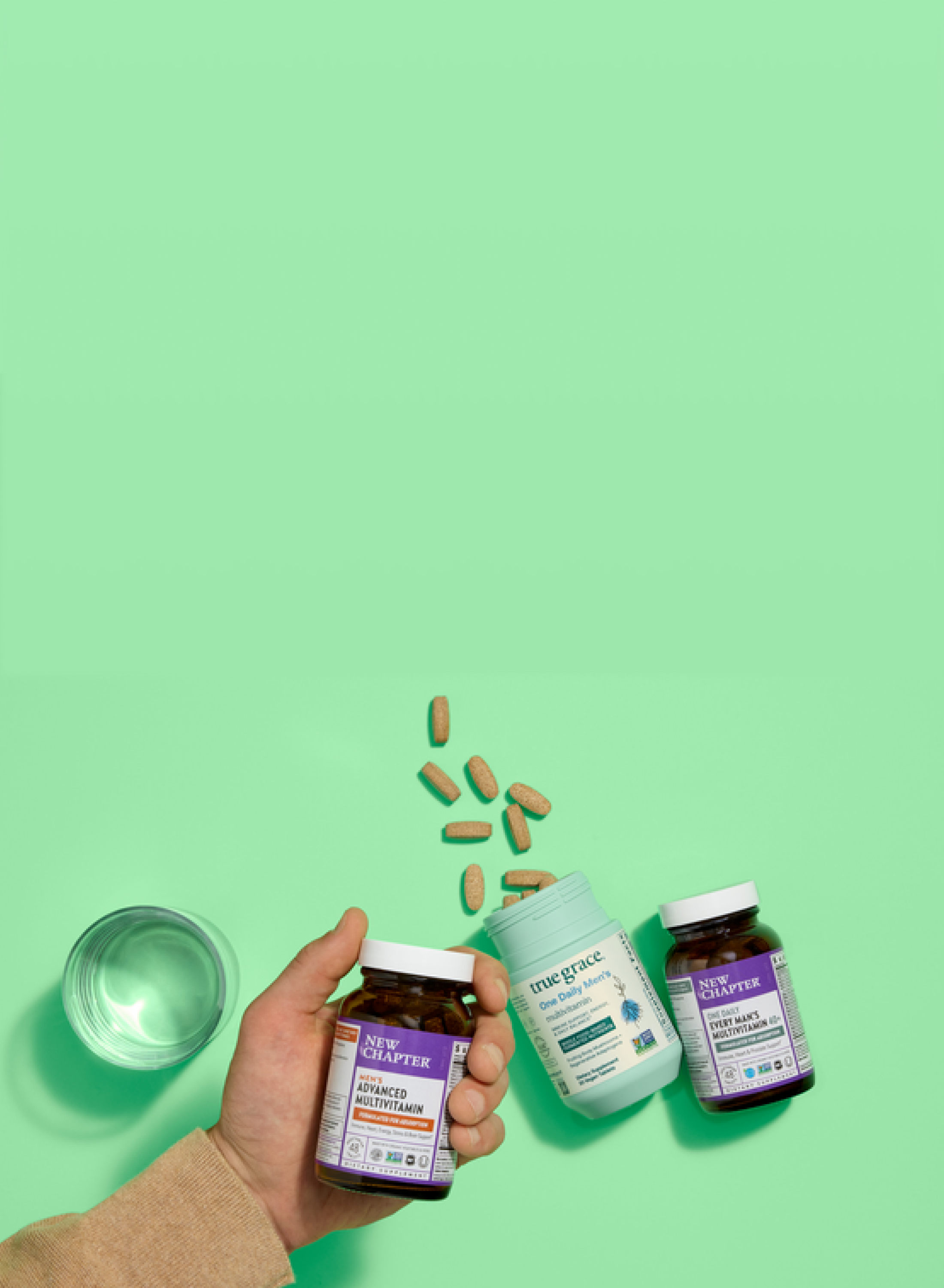 A hand holds a New Chapter multivitamin bottle next to another bottle and a True Grace container spilling capsules, with a glass of water, all on a mint green background.