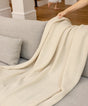 A person’s hand drapes a cream-colored, plant-dyed Adult Popcorn Blanket - Cream over a gray sofa.
