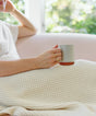 A person sitting on a sofa holds a mug while covered by a cream-colored, textured Adult Popcorn Blanket - Cream.