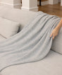 A hand holds the corner of a textured, light gray Adult Popcorn Blanket - Moonstone draped over a sofa.