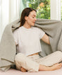 A person sits cross-legged while holding a large, grey, textured Adult Popcorn Blanket - Moonstone over their shoulders.