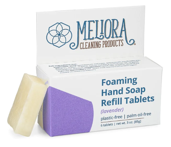 A box labeled Meliora Cleaning Products Foaming Hand Soap Refill Tablets (lavender) is open, showing a purple interior. One beige soap tablet is placed outside the box. The box notes it is plastic-free and palm oil-free.