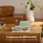 A tidy wooden table holds books, a basket, and a vase of flowers. Text reads: Mrs. Meyer's Multi-Surface Cleaner removes dirt and freshens surfaces.