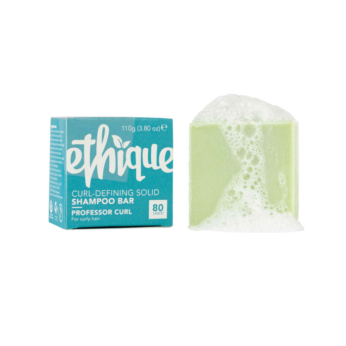 A green Ethique Curl-Defining Solid Shampoo Bar, partially covered in white suds, sits beside its turquoise and white packaging labeled “Curl-Defining Solid Shampoo Bar, Professor Curl, 80 uses, 110g.”.