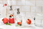 Glass soap dispensers with floral designs and silver pumps are on a white marble countertop next to pomegranates, a clear pitcher of water, and a glass, against a white tiled wall.