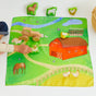 A child enjoys imaginative play on Sarahs Silks Play Map, a vibrant farm-themed mat inspired by Montessori principles. It features a barn, sheep, chickens, and a pond with soft toys scattered around and the child holding a plush sheep beside a woven basket.