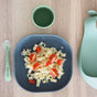 A gray bowl with pasta and halved cherry tomatoes on a wooden table, next to a green baby spoon, a green cup of water, and a matching green silicone bib.