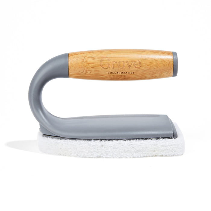 The Tub Handler + Build Up Banisher by Grove Co. features a curved handle of gray plastic and light wood, with a white pad for deep cleaning, casting a subtle shadow on a plain white background.