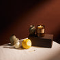 A lit P.F. Candle Co. Soy Wax Candle in an amber jar sits on a block near small ceramic vases, a yellow apple, and a matchstick, all arranged on a white cloth against a brown background. Warm, soft lighting enhances the cozy atmosphere.
