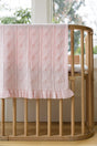 A light pink AU Baby Little Love Blanket - Peony in organic merino wool with a heart pattern and a ruffled edge draped over a wooden crib.