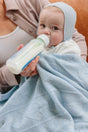 An adult holds a baby wearing a light blue bonnet and wrapped in an organic Cashmere Cloud Baby Blanket Gift Set - Hydrangea while drinking from a bottle.
