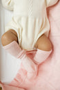 An infant in a cream-colored knit onesie and light pink socks lying on a Cashmere Cloud Baby Blanket Gift Set - Peony Pink with a pink pattern.