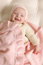 A smiling baby in a light pink bonnet and cream sweater, wrapped in a matching AU Baby Cashmere Cloud Baby Blanket Gift Set - Peony Pink.