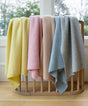 Four superfine merino waffle-knit blankets, including The Popcorn Blanket - Sky Blue, in yellow, pink, beige, and light blue draped over a wooden crib rail.