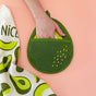 A hand slips into The Neighborgoods Pot Holder - Avocado, a playful oven mitt shaped like an avocado, resting on a peach surface next to a white and green avocado-patterned cloth.