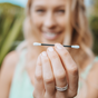 A woman with long blonde hair holds a reusable black cotton swab with white textured tips, smiling in an outdoor setting. The focus is on the swab and her hand, with a blurred background of greenery.